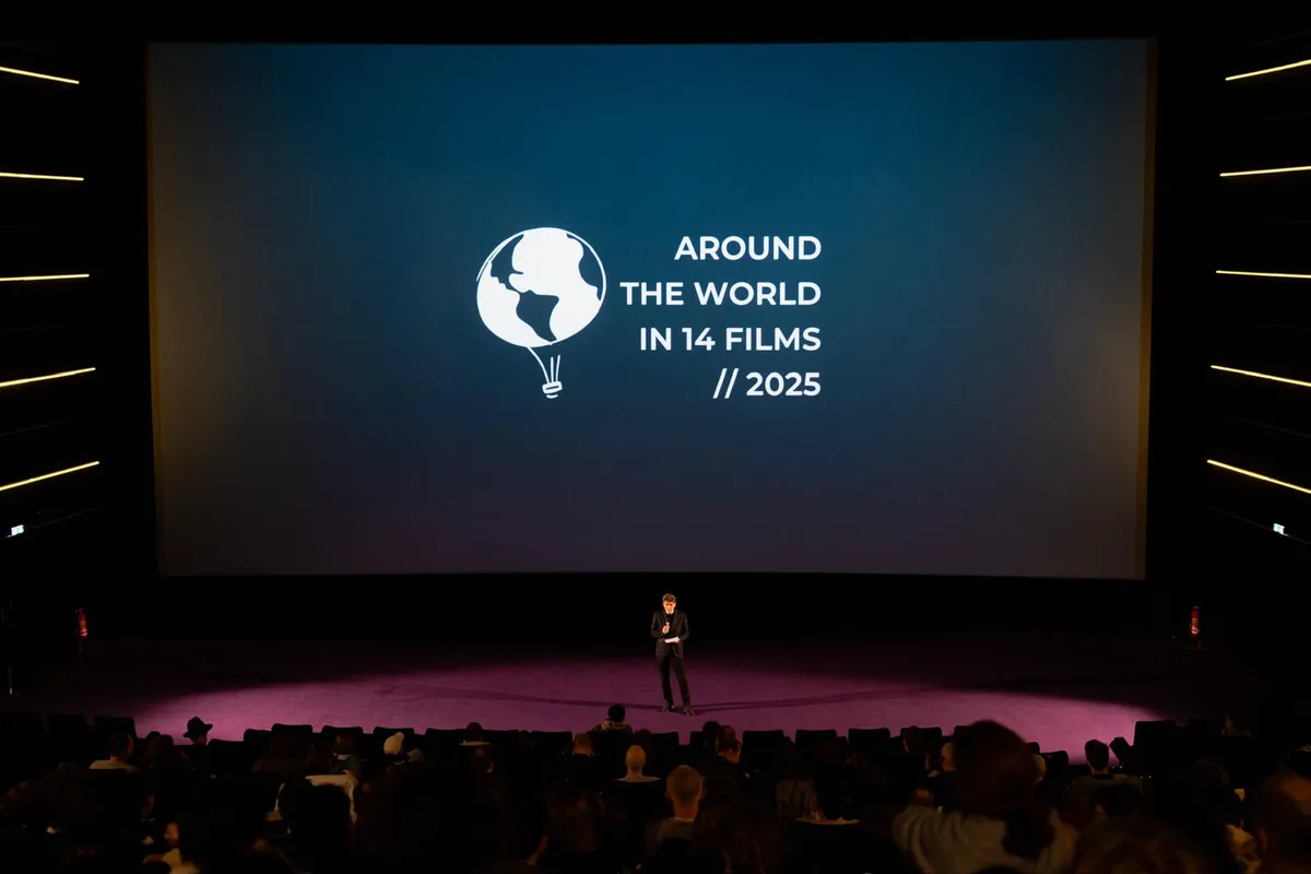 A full house, the festival logo with the year 2025 on the screen, a person speaking on stage in front of it